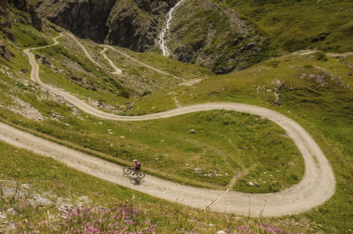 A lone biker pedals up a long, winding mountain trail.