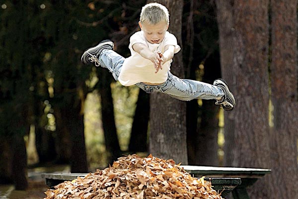 A boy jumping off a picnic table to bellyflop into a huge pile of leaves.