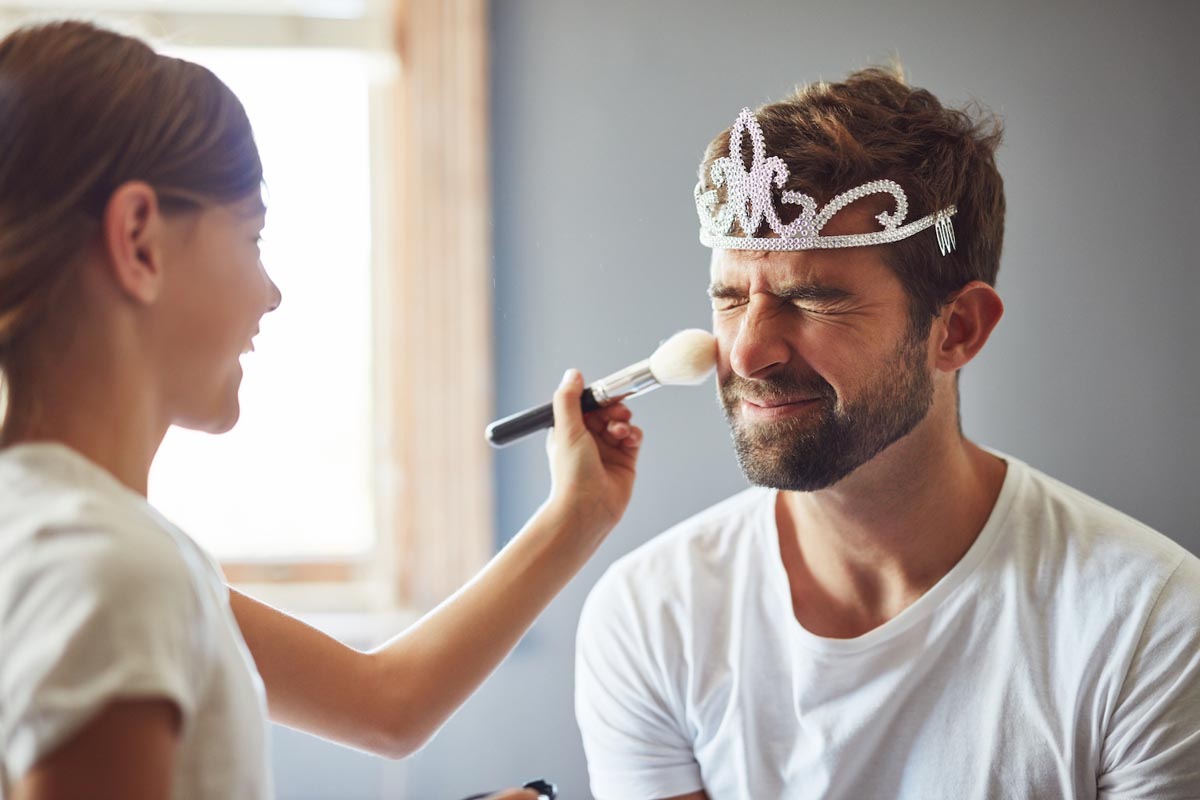 A father wearing a tiara and scrunching his face as his laughing daughter pretends to do his make-up.