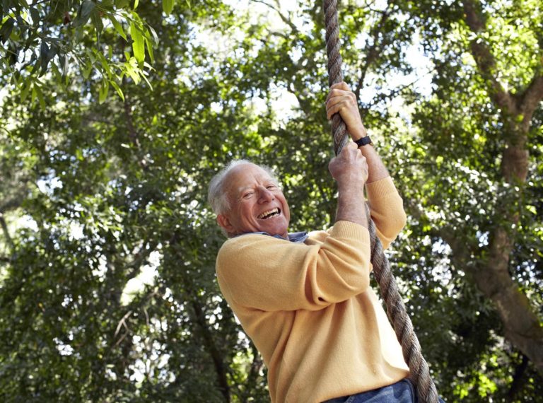 A laughing, gray-haired man in a yellow sweater swinging on a treehouse rope swing.