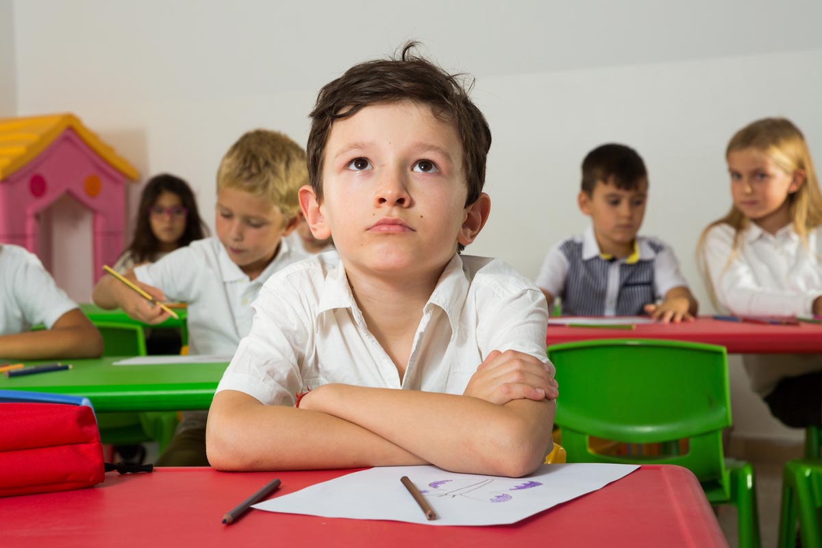 An unhappy kindergartener daydreaming at his school desk while his teacher helps classmates.