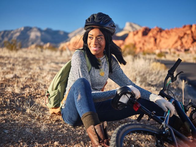 A smiling woman sitting next to a mountain trail with her bike, enjoying the view.